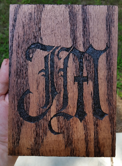 Picture of a stained slab of wood with the initials JM (in gothic font) carved into it.